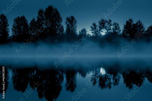 Night mystical scenery. Full moon through the tree branches, rising over the foggy river and its reflection in the still water. © stone36