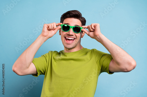 Photo of funky excited young man wear green t-shirt arms dark glasses smiling isolated blue color background