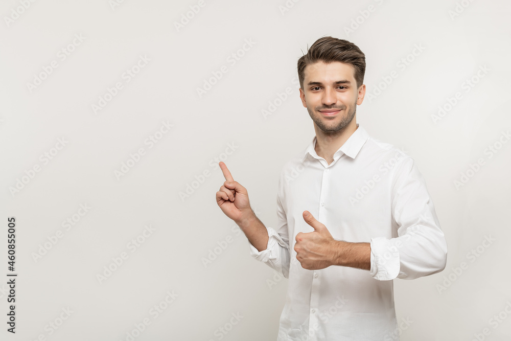 Young smiling man in in white shirt pointing left with his finger and showing thumb up isolated on white background. Young smiling man in in white shirt pointing left with his finger and showing thumb up isolated on white background.