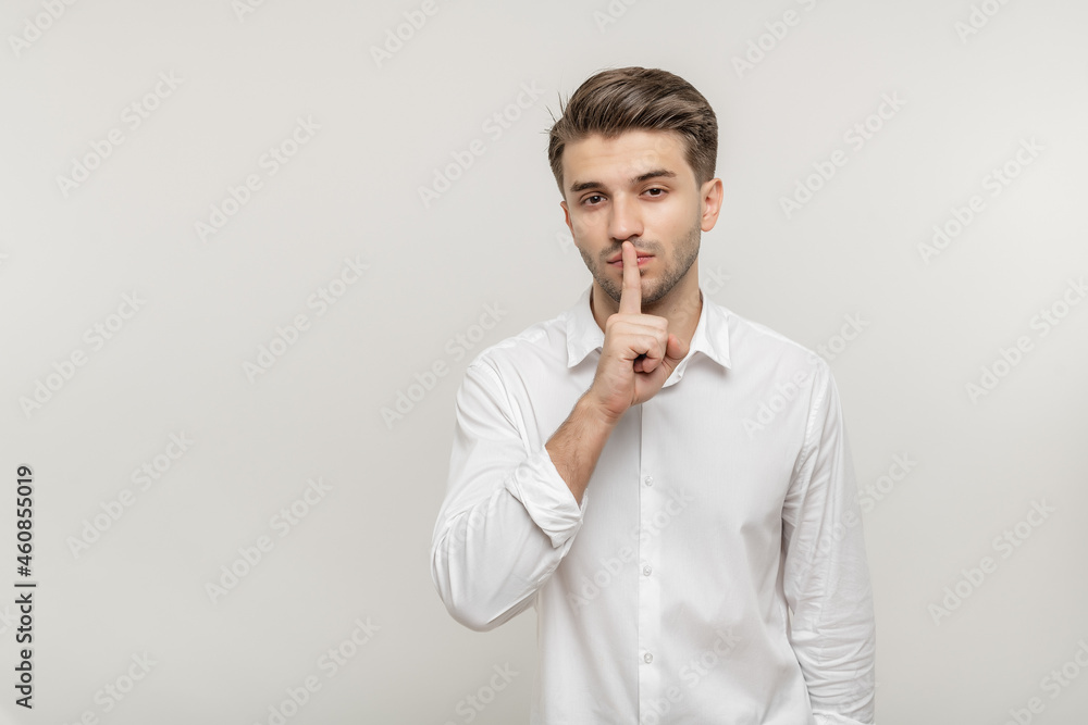 Young handsome man in white shirt makes gesture quietly, asks remain silent, gossips about something, looks mysteriously as tells secret, isolated on white background Young handsome man in white shirt makes gesture quietly, asks remain silent, gossips about something, looks mysteriously as tells secret, isolated on white background