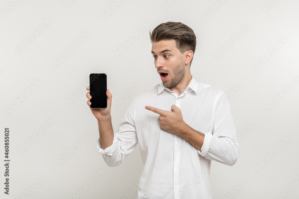 Young handsome happy man wearing white shirt, holding blank screen smartphone, pointing and watching at mobile, isolated over white background Young handsome happy man wearing white shirt, holding blank screen smartphone, pointing and watching at mobile, isolated over white background