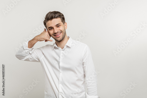 Smiling young unshaven business man in light shirt posing isolated on white wall background. Achievement career wealth business concept. Mock up copy space. Doing cellphone gesture © Anastasia