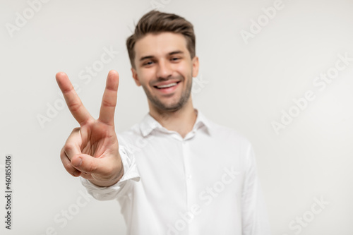 Young happy man with beard wearing white shirt standing over white background smiling and showing two fingers on camera hand doing victory sign. Number two. focus on hand
