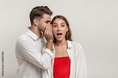 Image of handsome young man whispering secret or interesting gossip to shocked beautiful woman in her ear isolated over white background