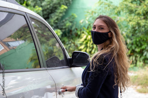 Brazilian woman using a black mask opening the door of a white car in the garage of her house. Background garden