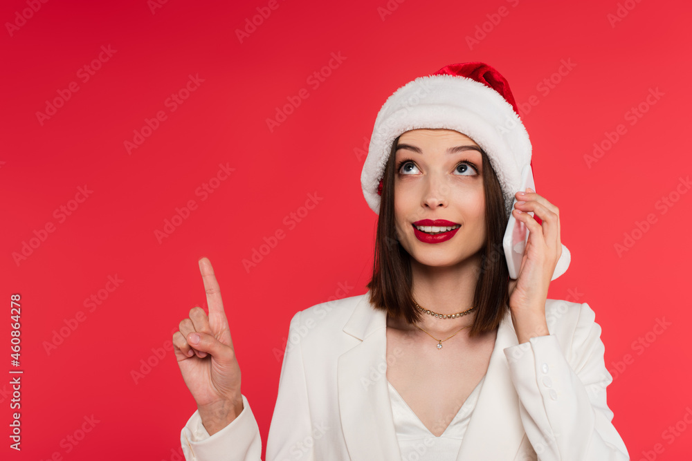 Smiling woman with red lips and santa hat talking on smartphone and pointing with finger isolated on red Smiling woman with red lips and santa hat talking on smartphone and pointing with finger isolated on red