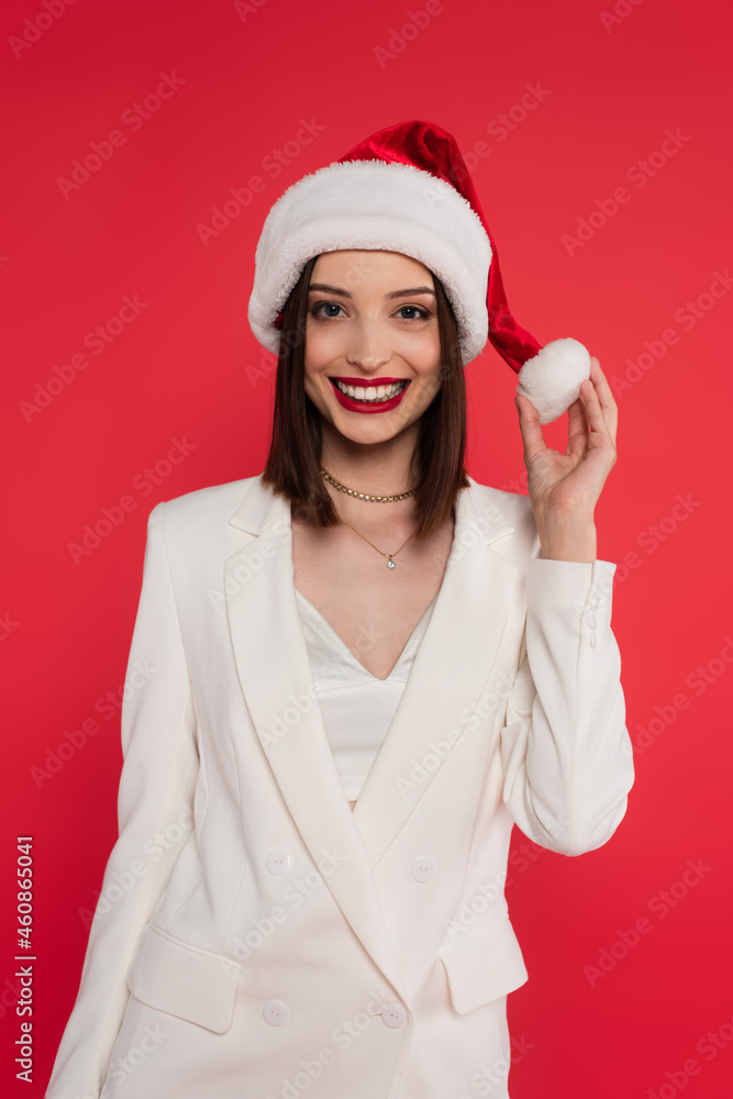 Positive woman in white jacket holding santa hat isolated on red Positive woman in white jacket holding santa hat isolated on red
