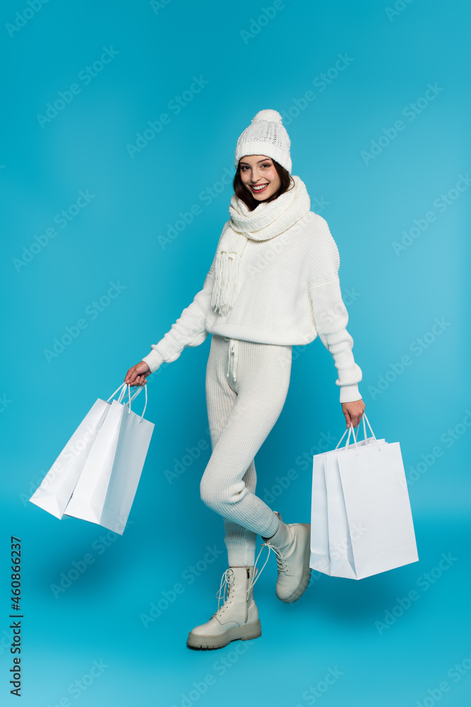 Full length of smiling woman in white warm clothes holding shopping bags on blue background Full length of smiling woman in white warm clothes holding shopping bags on blue background