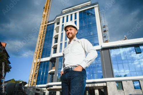 Successful engineer or architect, crane and building construction at backgrpound. Joyous businessman with wearing helmet.