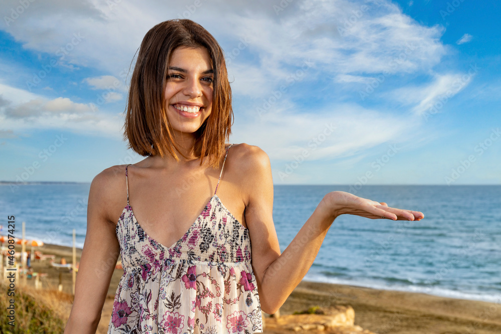 Smiling girl raising up her left hand to show a potential copy space on the right. Bright seaside landscape in the background for a summer concept.
