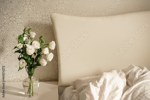 white flowers in a transparent glass vase stand on a bedside table by a bed with a large white headboard. bedroom interior. modern room
