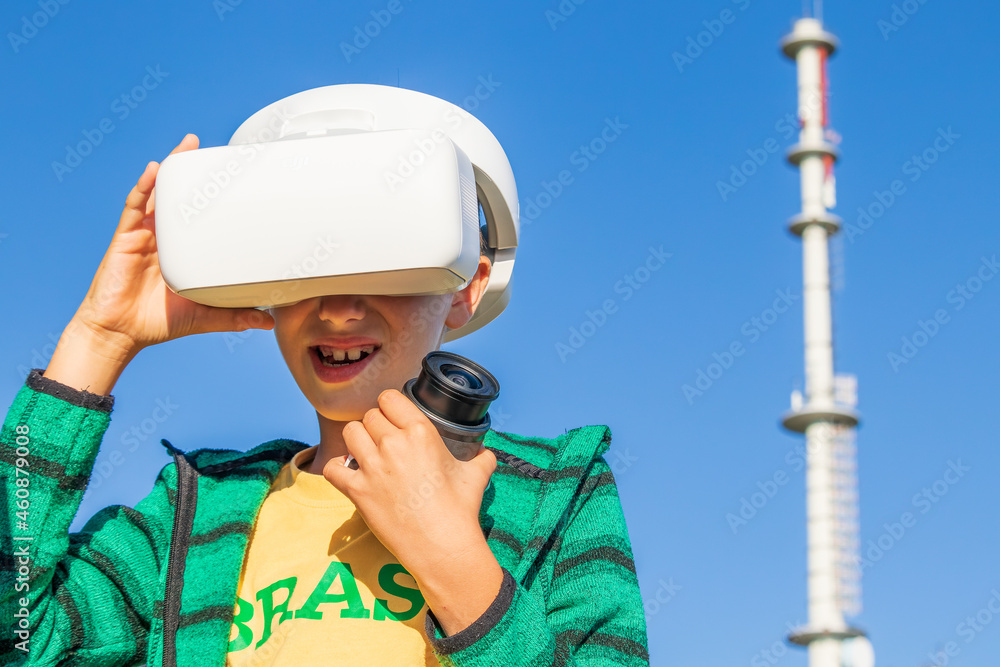 Obraz premium Child wearing virtual reality glasses and holding the camera in the left hand, in the background visible blue clear sky and mobile telephony relay