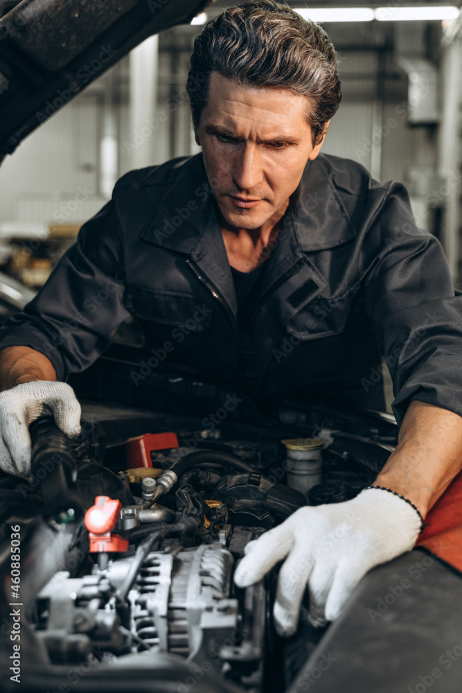 Waist up portrait image of senior serious mechanic in uniform standing near the opened car while working in auto service. Car repair and maintenance concept Waist up portrait image of senior serious mechanic in uniform standing near the opened car while working in auto service. Car repair and maintenance concept