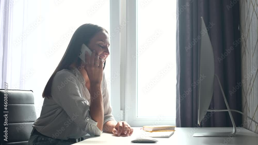 Smiling young woman with laptop talking on mobile phone sitting at the desk at home. Cheerful female freelancer talking on the smartphone and using PC, check some information online, making schedule Smiling young woman with laptop talking on mobile phone sitting at the desk at home. Cheerful female freelancer talking on the smartphone and using PC, check some information online, making schedule