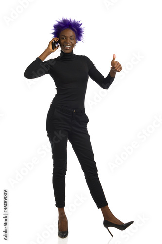 Relaxed black woman is standing, talking on the telephone and showing thumb up. Front view, full length, isolated.