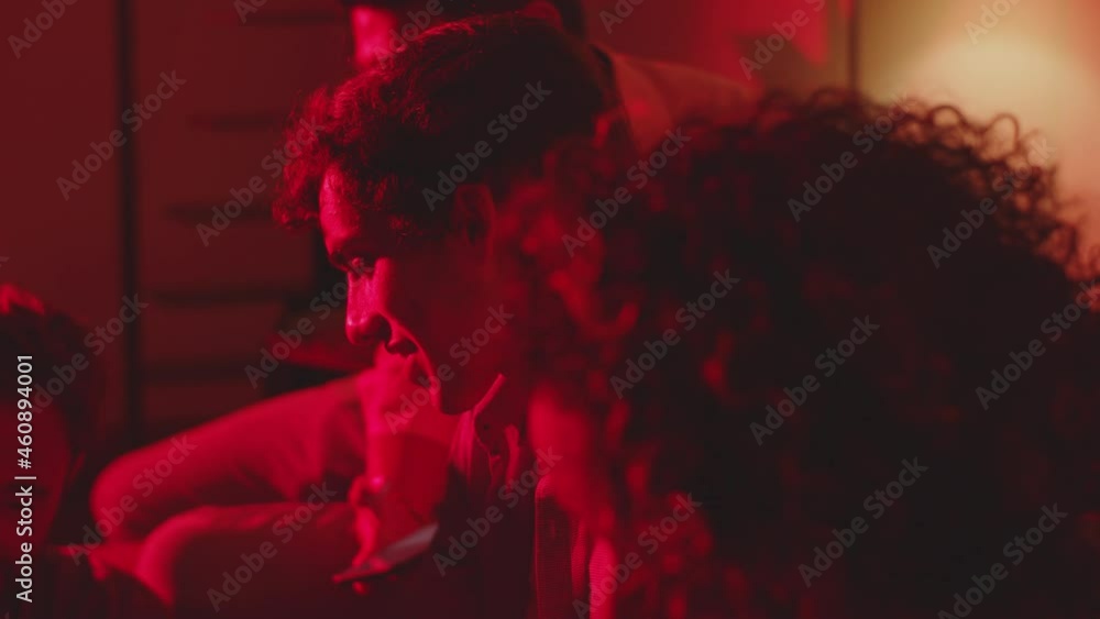 Group of friends talking in dark room with warm red light. Side view of cheerful men and women having party on weekend and enjoying time spending together. Home party, friendship concept Group of friends talking in dark room with warm red light. Side view of cheerful men and women having party on weekend and enjoying time spending together. Home party, friendship concept