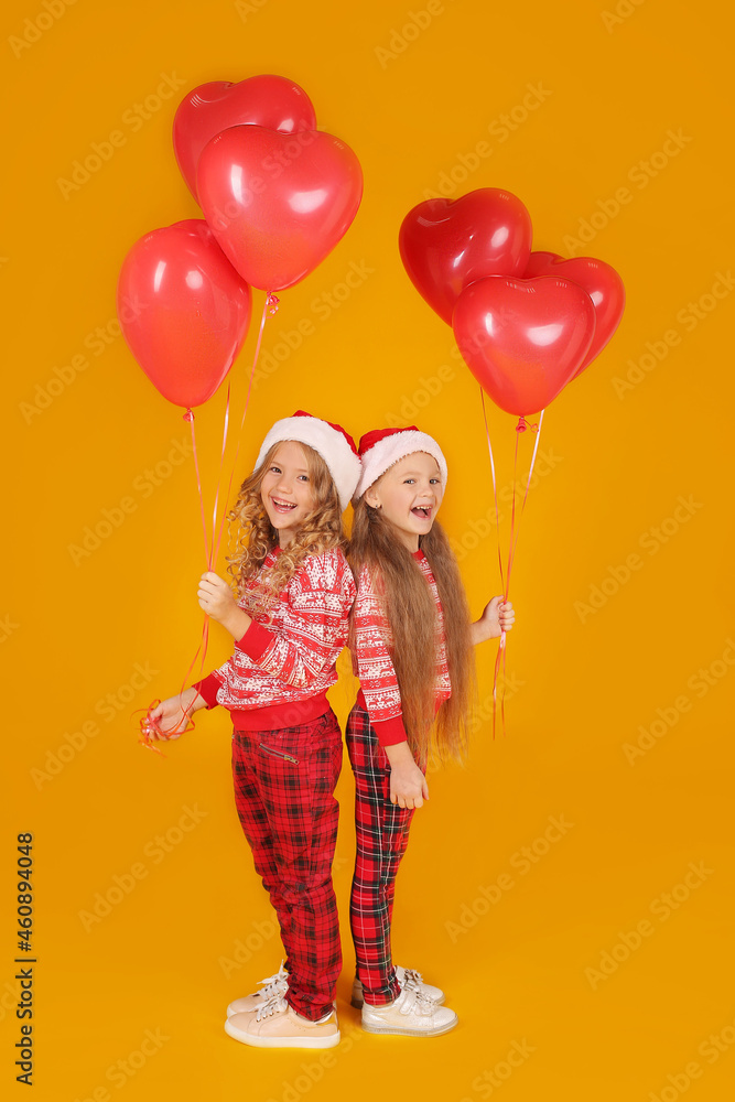 little beautiful happy blonde girls in a New Year's jumper in plaid trousers and Santa Claus hats are holding red balloons on a yellow background little beautiful happy blonde girls in a New Year's jumper in plaid trousers and Santa Claus hats are holding red balloons on a yellow background