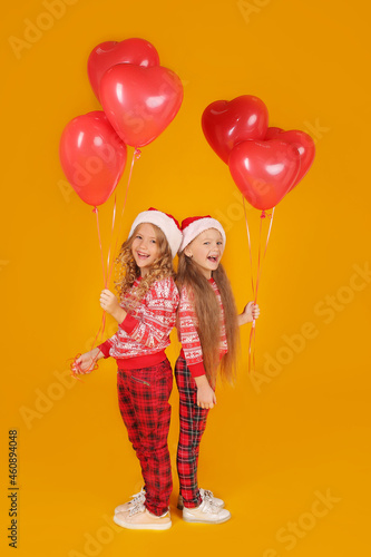 little beautiful happy blonde girls in a New Year's jumper in plaid trousers and Santa Claus hats are holding red balloons on a yellow background