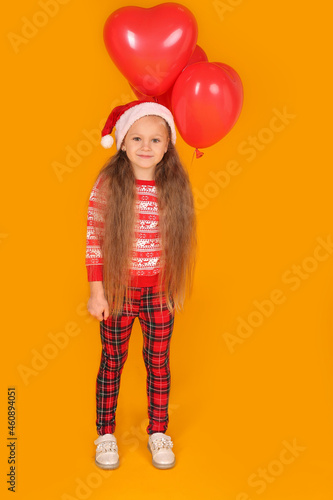 a little beautiful happy blonde girl in a New Year's jumper and plaid trousers with a Santa Claus hat holding red balloons on a yellow background