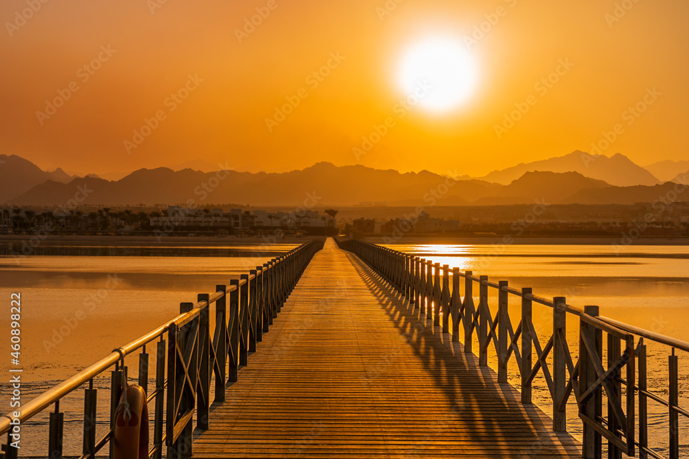 Picturesque bright orange sunset over silhouette of desert mountains of Egypt and coastline with reflection in sea water and wooden bridge road leading to sunlight on summer evening at vacation