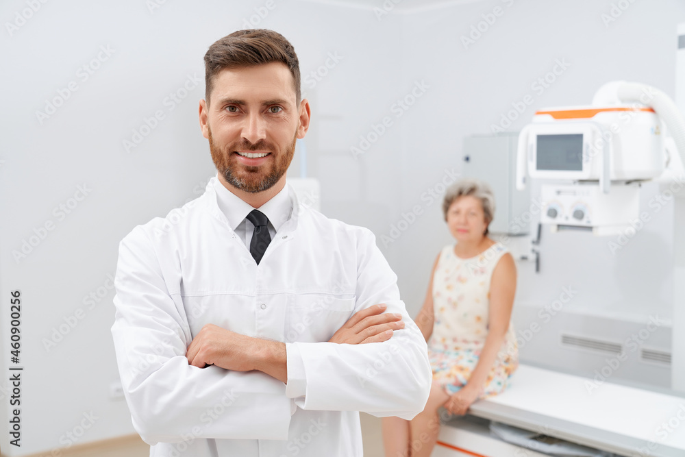 Front view of confident doctor in white uniform with crossed hands after success day after diagnostic. Professional radiologist and old female patient on blur background in modern clinic. Front view of confident doctor in white uniform with crossed hands after success day after diagnostic. Professional radiologist and old female patient on blur background in modern clinic.