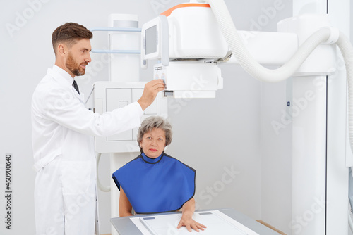 Front view of grey haired woman smiling, sitting at cabinet of ultra diagnostic, smiling at camera. Professional radiologist press buttons preparing usg above hand of patient. Concept of medicine.