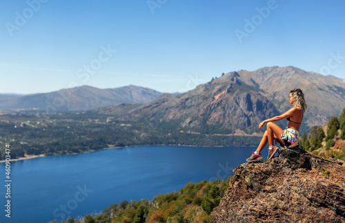 young woman sitting on a rock on top of a mountain looking at the landscape of mountains and blue lake