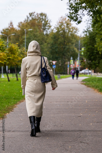a girl in a fashionable beige autumn coat, boots with a fashionable bag walks along a path in a city park © Андрей Знаменский