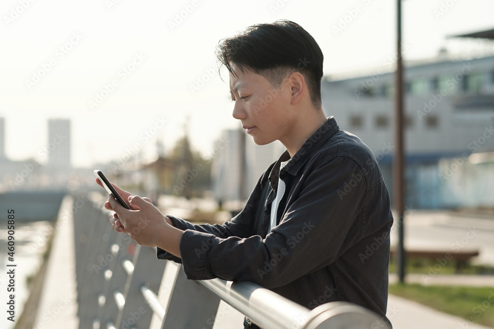 Young serious businesswoman looking at smartphone screen while standing by riverside Young serious businesswoman looking at smartphone screen while standing by riverside