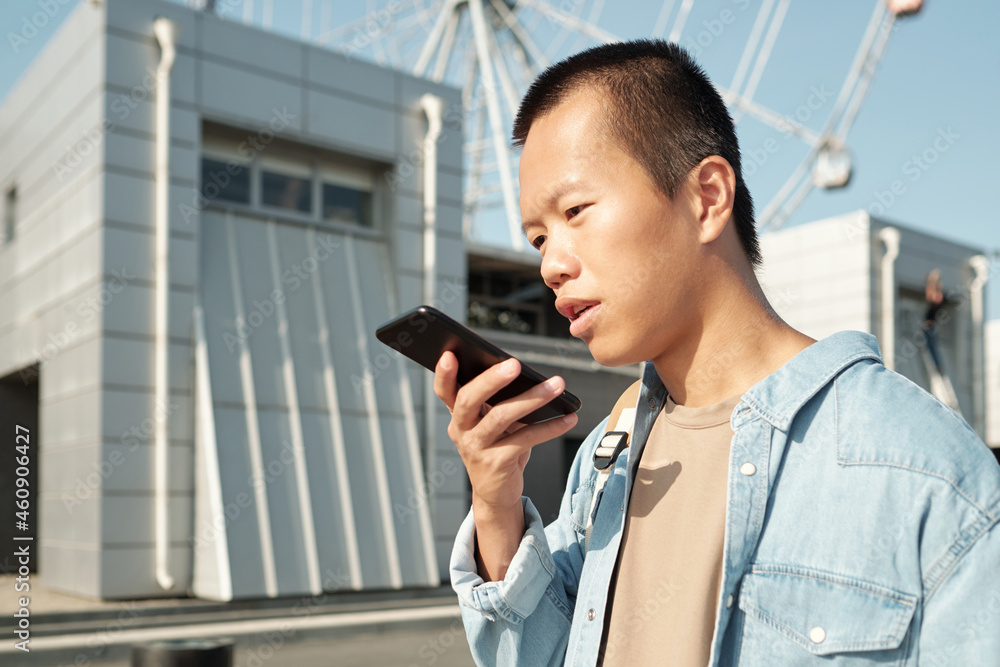 Fototapeta premium Young man of Asian ethnicity recording voice message against modern architecture