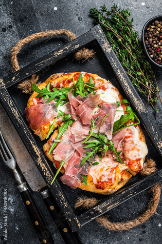 Italian Pizza with prosciutto parma ham, arugula salad and cheese in a rustic wooden tray. Black background. Top view