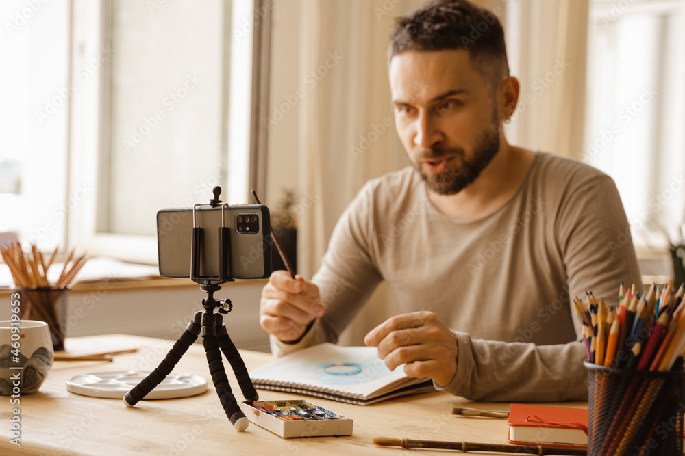 Portrait of Mid-adult artist male. Dark haired man smiling happy making online art class for students at studio. Holding watercolor drawing in his hand. Concept zoom Remote learning. Real emotions