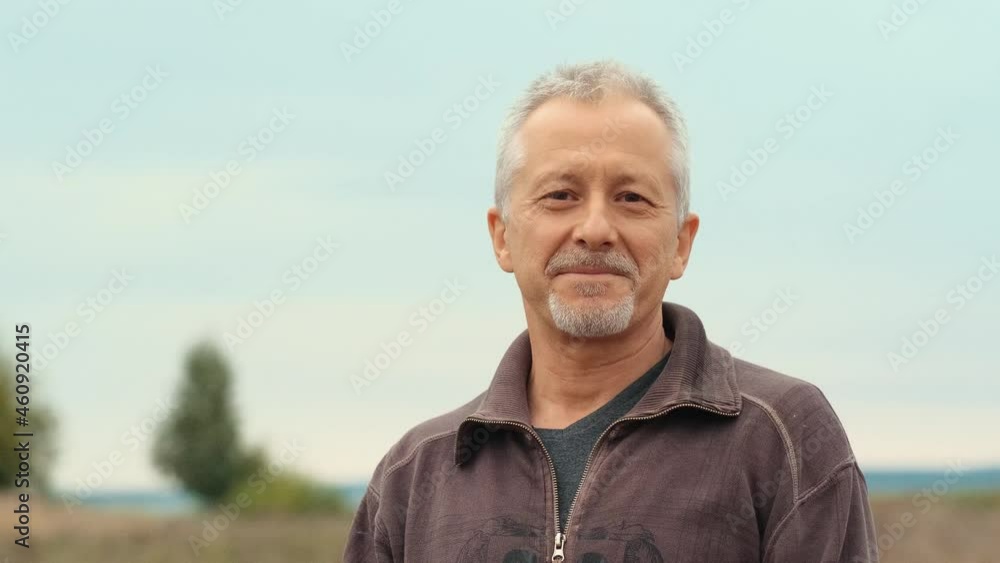 An elderly gray-haired happy man with a beard looks into the camera and smiles while standing in nature on a summer or autumn evening. An elderly gray-haired happy man with a beard looks into the camera and smiles while standing in nature on a summer or autumn evening.