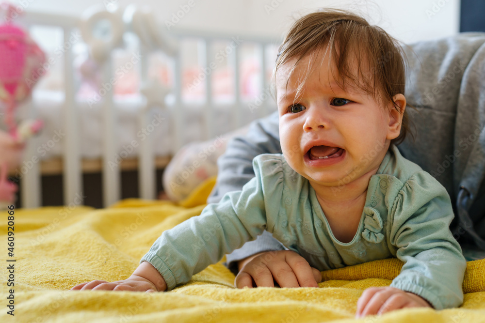 Front view on unhappy small caucasian baby lying on the bed crying by her mother due cramps or frustration at home in day copy space childhood growing up family concept