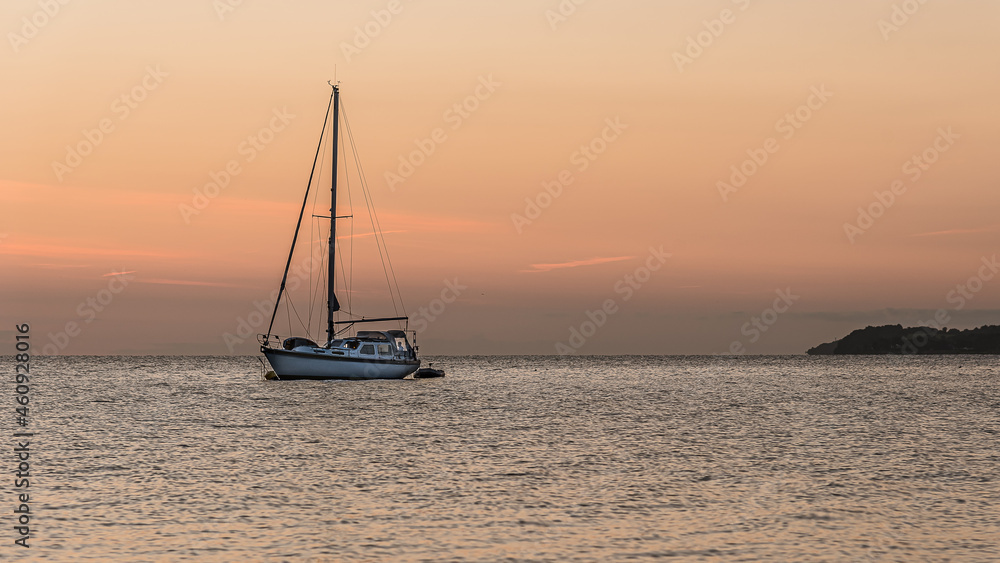 a sailboat is at anchor at sunset and a man is standing in a window in the stern