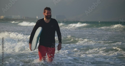 Portrait of a handsome senior sportsman at the age carrying a surfboard on the beach. A retired athlete enters the sea, goes to ride on the waves on a surfboard.