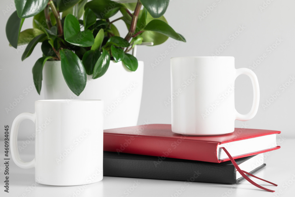 Two mugs mockup with notebook and green plant in pot on white table. front view Two mugs mockup with notebook and green plant in pot on white table. front view