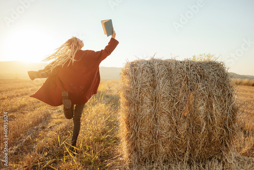 Cheerful young woman, dancing and jumping while reading an exciting book in the field outside