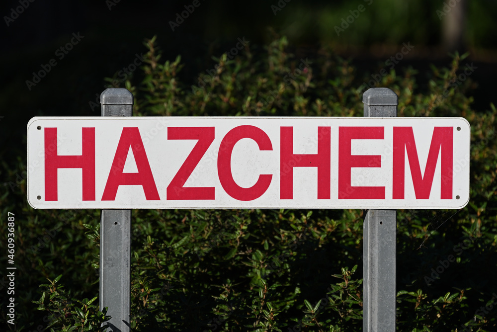 A white hazchem sign with bright red lettering, held up by two steel posts. The dirty sign sits in front of a dark green bush, and has a spiderweb attached to its bottom right A white hazchem sign with bright red lettering, held up by two steel posts. The dirty sign sits in front of a dark green bush, and has a spiderweb attached to its bottom right