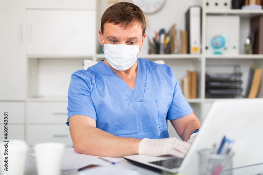 Confident male doctor in blue medical uniform, mask and gloves working on laptop in clinic office
