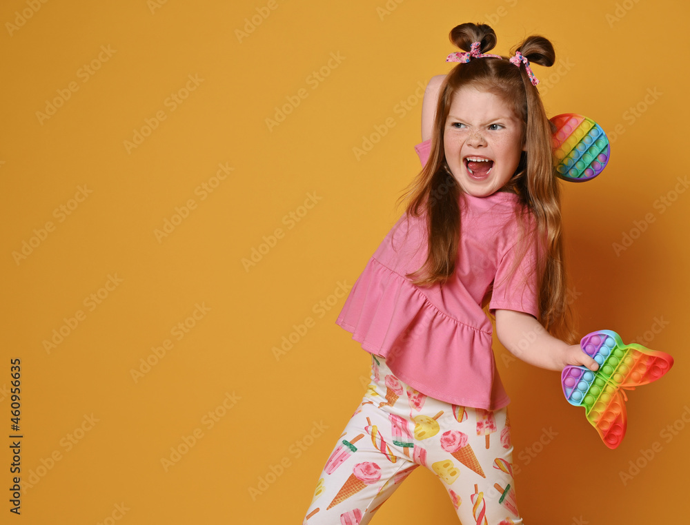 Screaming, spoiled redhead kid girl in colorful clothes is going to throw away two sensory rainbow color toys - round and butterfly shape pop it over background with copy space