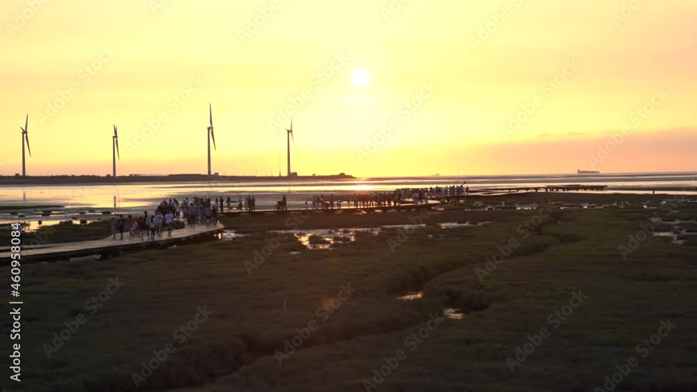 Vivid bright orange sunset landscape shot of tourists strolling on wooden walkway platform at beautiful Gaomei wetlands preservation area with wind power plant in the background in Taichung, Taiwan. Vivid bright orange sunset landscape shot of tourists strolling on wooden walkway platform at beautiful Gaomei wetlands preservation area with wind power plant in the background in Taichung, Taiwan.