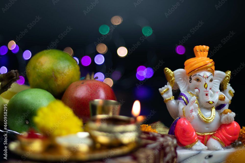 Colorful Statue Of Hindu God Ganesha Ganpati Bappa Morya Sitting On Golden Asana With Fruits, Flowers, Lamp And Blur Bokeh For Diwali Puja New Year Deepawali Ganesh Chaturthi Or Shubh Deepavali Pooja