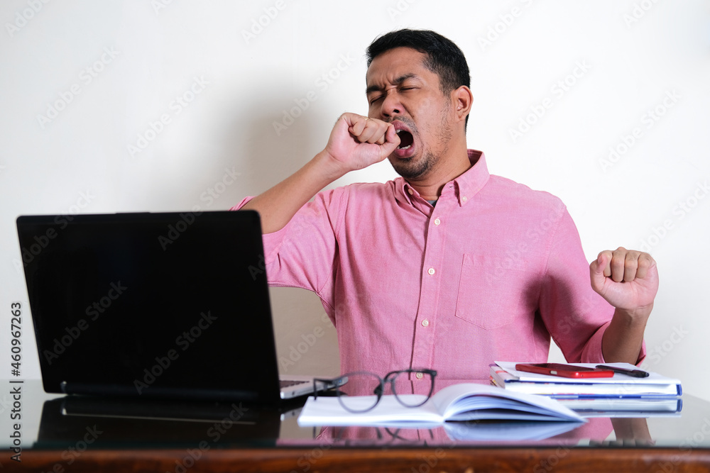 Adult Asian man showing tired gesture in front of his working laptop Adult Asian man showing tired gesture in front of his working laptop