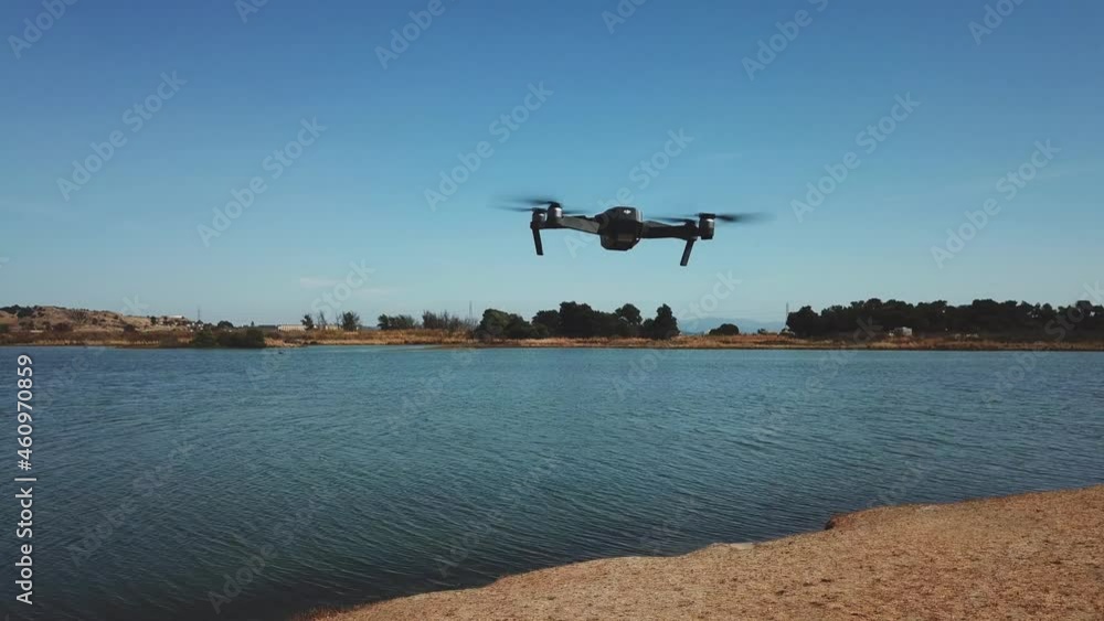 Quadcopter drone hovers head height lakeside in light breeze on blue sky day. Quadcopter drone hovers head height lakeside in light breeze on blue sky day.