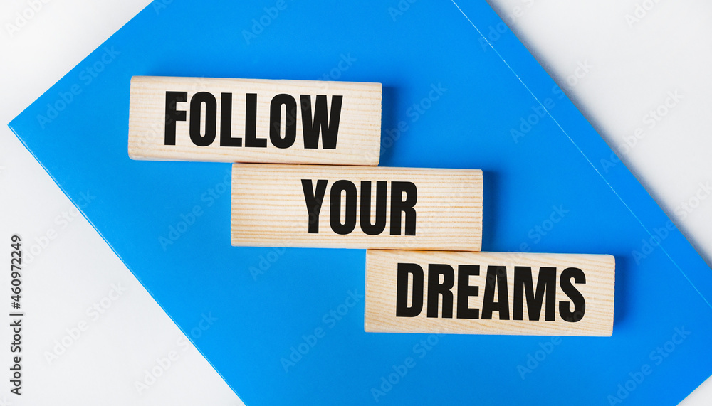 There is a blue notebook on a light gray background. Above are three wooden blocks with the words FOLLOW YOUR DREAMS