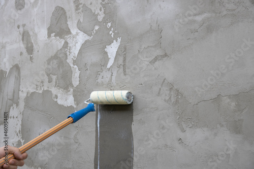 A worker applies the primer to the wall with a long-handled roller. A man's hand rolls a primer onto a gray wall. Wet footprint on a plastered wall. House facade renovation concept.