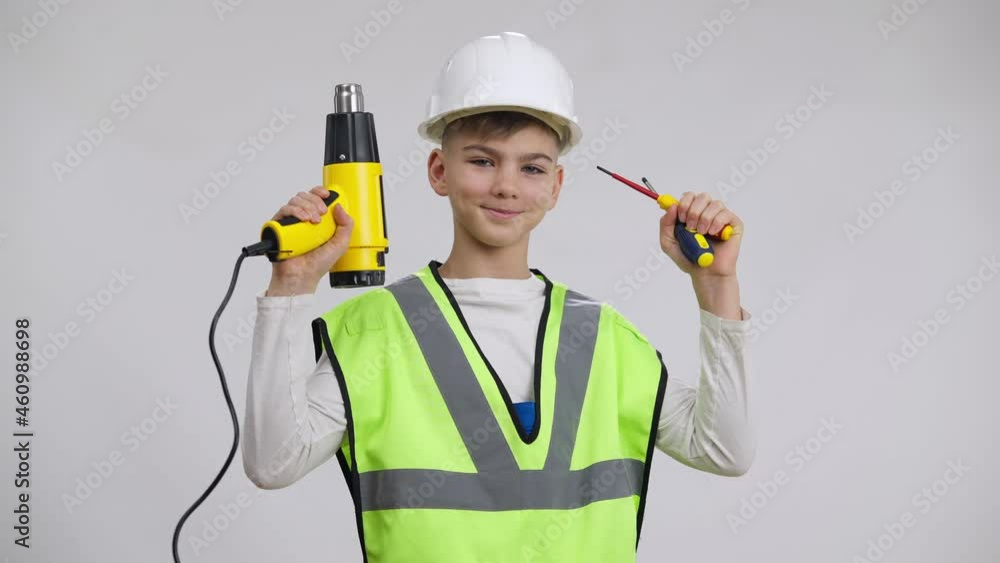 Cute Caucasian boy in hard hat raising tools smiling posing at white background. Confident happy kid choosing profession of repairman looking at camera holding screwdrivers and perforator Cute Caucasian boy in hard hat raising tools smiling posing at white background. Confident happy kid choosing profession of repairman looking at camera holding screwdrivers and perforator