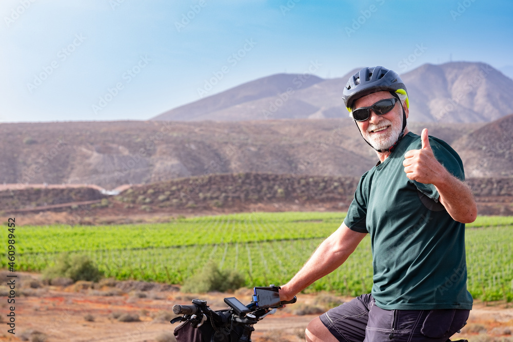 Senior man on his electro bike in outdoor excursion gesturing positive sign with tumb up. Sporty helmet and sunglasses. Green vineyard and mountain in background