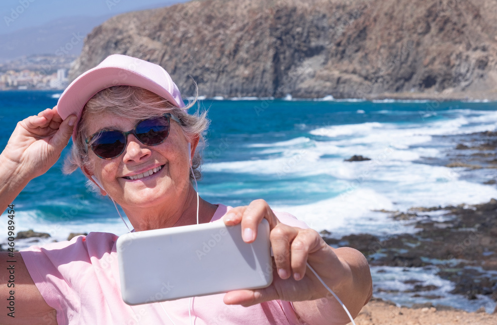 Attractive elderly woman holding mobile phone taking selfie at sea in windy day. Senior people enjoying holiday and beauty in nature, mountain, beach and waves on background